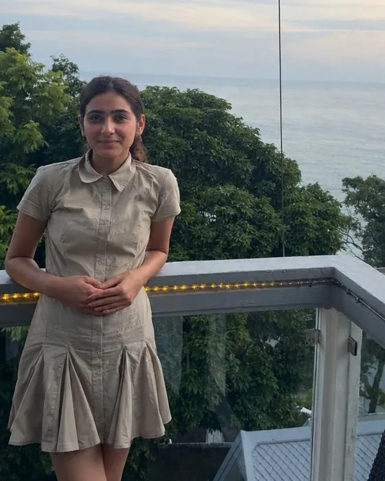 Jessica in a beige dress leaning on a balcony railing with the sea and trees behind her